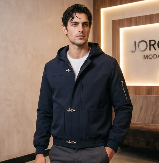 Chaqueta bomber para hombre con capucha, confeccionada en tejido liso disponible en color negro y azul marino, con bolsillos delanteros, bolsillo utilitario con cremallera plateada en la manga, bajo elástico fruncido y cierres metálicos tipo mosquetón al frente