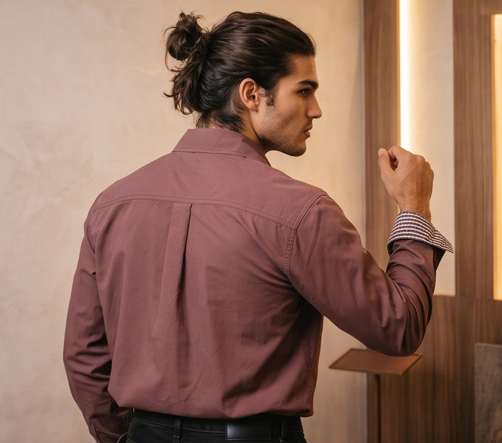 Camisa casual de hombre confeccionada en algodón liso, de corte holgado y manga larga, con cuello de solapa camisero abotonado al frente y detalle estampado en el borde interno de los puños y cuello, mostrada de frente y de espaldas en tonos café rojizo y gris oscuro azulado.