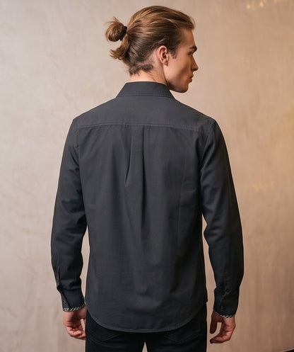 Camisa casual de hombre confeccionada en algodón liso, de corte holgado y manga larga, con cuello de solapa camisero abotonado al frente y detalle estampado en el borde interno de los puños y cuello, mostrada de frente y de espaldas en tonos café rojizo y gris oscuro azulado.
