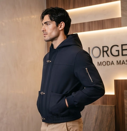 Chaqueta bomber para hombre con capucha, confeccionada en tejido liso disponible en color negro y azul marino, con bolsillos delanteros, bolsillo utilitario con cremallera plateada en la manga, bajo elástico fruncido y cierres metálicos tipo mosquetón al frente