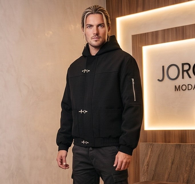 Chaqueta bomber para hombre con capucha, confeccionada en tejido liso disponible en color negro y azul marino, con bolsillos delanteros, bolsillo utilitario con cremallera plateada en la manga, bajo elástico fruncido y cierres metálicos tipo mosquetón al frente