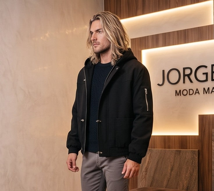 Chaqueta bomber para hombre con capucha, confeccionada en tejido liso disponible en color negro y azul marino, con bolsillos delanteros, bolsillo utilitario con cremallera plateada en la manga, bajo elástico fruncido y cierres metálicos tipo mosquetón al frente