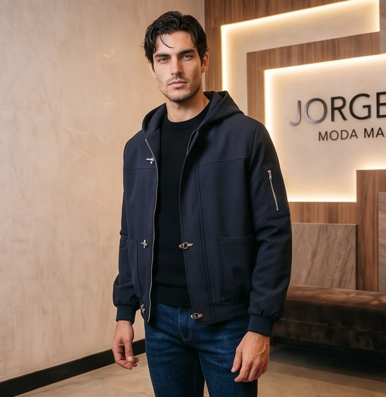 Chaqueta bomber para hombre con capucha, confeccionada en tejido liso disponible en color negro y azul marino, con bolsillos delanteros, bolsillo utilitario con cremallera plateada en la manga, bajo elástico fruncido y cierres metálicos tipo mosquetón al frente