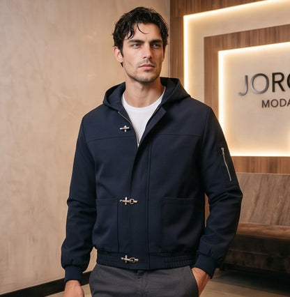Chaqueta bomber para hombre con capucha, confeccionada en tejido liso disponible en color negro y azul marino, con bolsillos delanteros, bolsillo utilitario con cremallera plateada en la manga, bajo elástico fruncido y cierres metálicos tipo mosquetón al frente
