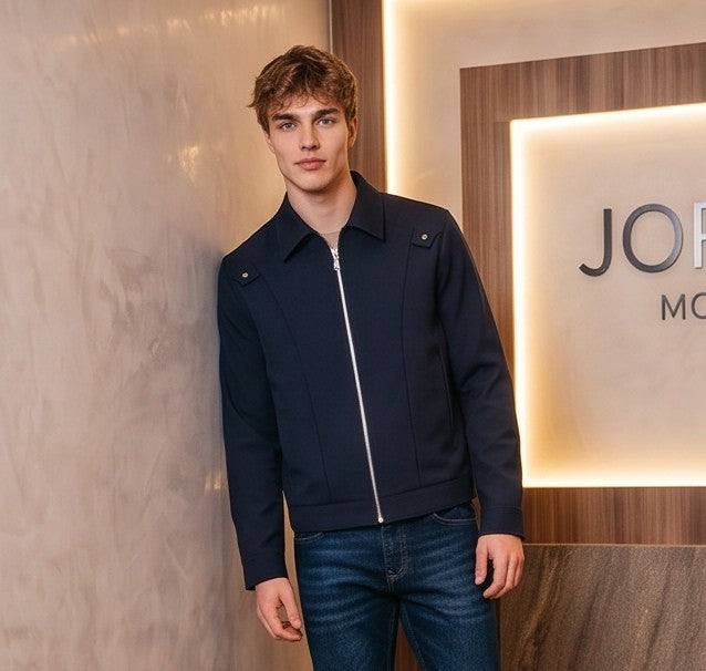 Chaqueta entallada slim fit para hombre disponible en color negro y azul marino, confeccionada en tejido liso estructurado con cuello camisero, cremallera metálica frontal central, puños con cierre a presión y detalles decorativos de solapa en los hombros sobre fondo de estudio minimalista