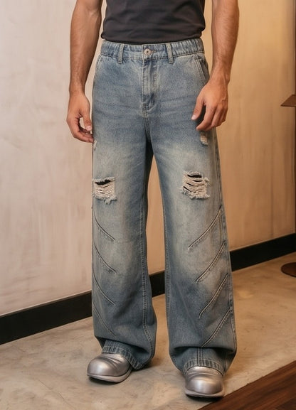 Pantalón jean ancho estilo urbano streetwear en denim 100% algodón con efecto lavado en tonos azul y negro desgastado, tiro medio-alto, cintura semielástica con botón y cremallera, roturas desgastadas en rodillas y bordados diagonales decorativos en las piernas