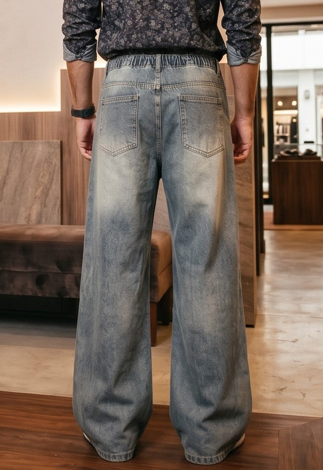 Pantalón jean ancho estilo urbano streetwear en denim 100% algodón con efecto lavado en tonos azul y negro desgastado, tiro medio-alto, cintura semielástica con botón y cremallera, roturas desgastadas en rodillas y bordados diagonales decorativos en las piernas